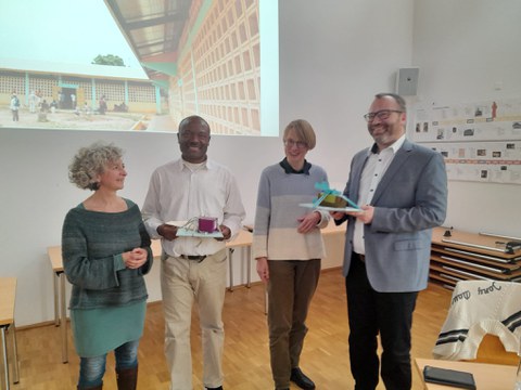 Vier Personen stehen in einem hellen Seminarraum nebeneinander und lächeln in die Kamera. Im Hintergrund sind Tische, Wandplakate und eine große Projektion zu sehen.