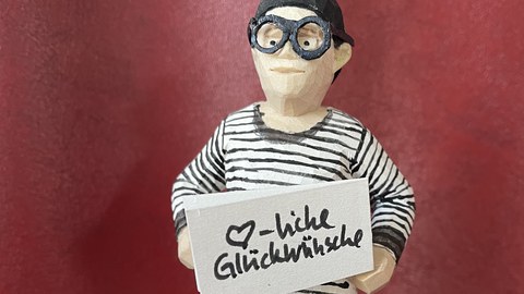 Eine Holzfigur die wie Anja Besand aussieht hält ein Schild mit der Aufschrift Herzliche Glückwünsche 