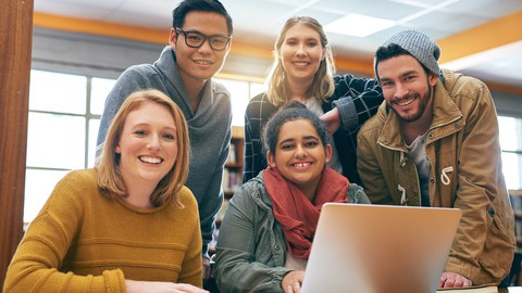 Foto einer Gruppe von 5 Studenten, die gemeinsam in der Bibliothek zusammen lernen und fröhlich in die Kamera lächeln