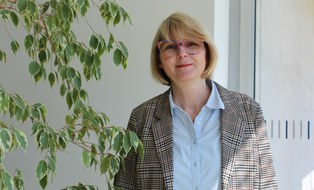 Caroline Friedrich ist eine weiße Frau mittleren Alters, die vor einer weißen Wand steht. Sie trägt ihre Haare in einem kurzen, blonden Bob mit Pony und trägt eine bunte Brille. Über einer hellblauen Bluse trägt sie einen braunkarierten Blazer. Auf dem Bild ragen links Blätter einer Pflanze ins Bild. Caroline Friedrich lächelt freundlich in die Kamera.