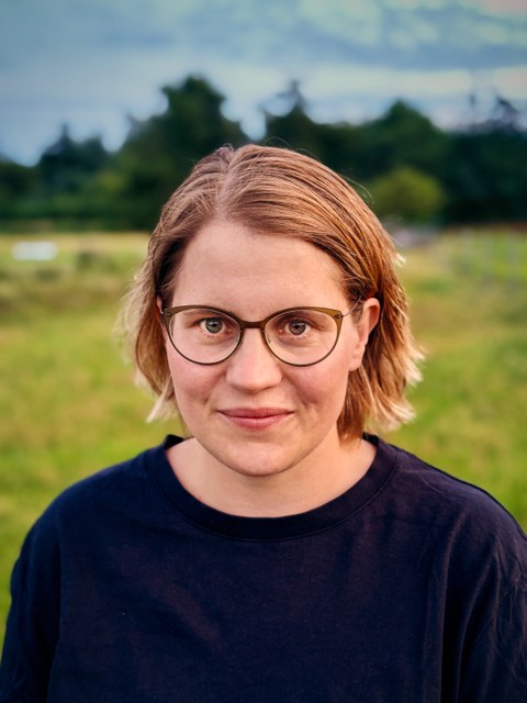 Porträtfoto von Anne Piezunka: Sie blickt frontal in die Kamera, lächelt dabei, trägt eine Brille und ein dunkles Oberteil. Sie steht auf einer Wiese, im Hintergrund sind unscharf Bäume zu erkennen.