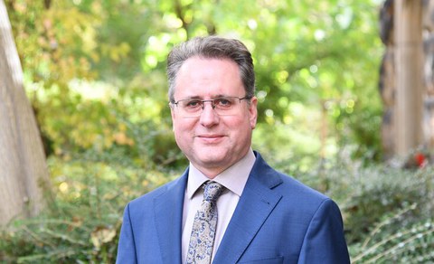 Porträtfoto von Jörg Ganzenmüller: Er steht im Anzug und mit Krawatte vor Bäumen und Büschen mit grünen Blättern. Er trägt eine Brille und lächelt in die Kamera.