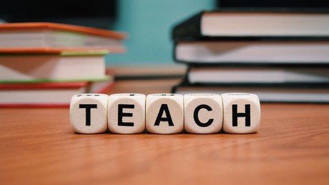 Das Bild zeigt fünf Würfel mit Buchstaben, die das Wort teach bilden. Im Hintergrund sind Bücher zu sehen.
