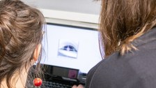 Zwei Frauen schauen auf einen Bildschirm, auf dem ein Auge mit Fadenkreuz zu sehen ist