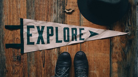 Zu sehen ist ein dreieckiges weißes Banner auf einem hölzernen Fußboden, bedruckt mit dem Wort Explore. Unter dem Banner sind ein paar Schuhe zu sehen, daneben ein Hut. 