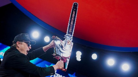 Elon Musk ist auf dem Bild zu sehen, wie er eine Kettensäge in die Höhe hält