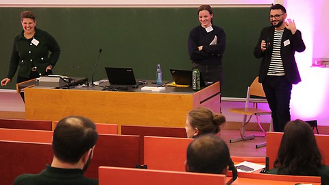 3 Personen vor einer Tafel die lachen und reden und präsentieren