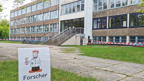 Das Unischulgebäude, ein DDR-Plattenbau, ist schräg von vorn zu sehen. Davor hängt an rot-weißem Flatterband ein Schild mit einer gezeichneten Comicfigur eines Forschers. Darunter steht "Gruppe Forscher B". Im Hintergrund ist der Schulhof mit rot-weißem Flatterband in verschiedene Wartezonen abgteilt. 