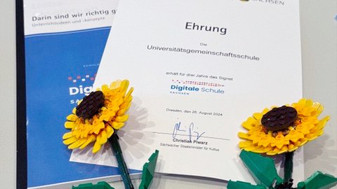 Die Urkunde der Auszeichnung "Digitale Schule Sachsen" für die Universitätsgemeinschaftsschule Dresden und ein Heft zum Netzwerk Digitale Schule Sachsen. Darauf zwei Legeo-Sonnenblumen