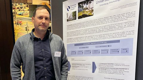 Ein Mann mit grauem Jacket steht vor einem wissenschaftlichen Poster.