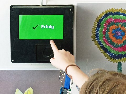 An einer Wand mit bunten Kinderbildern hängt ein schwarzer Kasten mit grünem Bildschirm. Auf diesem erscheint ein schwarzes Häkchen und der weiße Schriftzug "Erfolg". Eine Kinderhand hält einen Chip für die Anmeldung in der Hand.