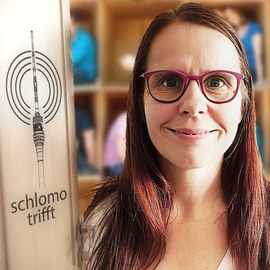 Das logo der ColoRadio-Sendung Schlomo trifft auf einem Türrahmen, daneben lächelt eine Frau mit Brille frontal in die Kamera. Im Hintergrund ein großes Regal mit Jugendlichen in großen Fächern.