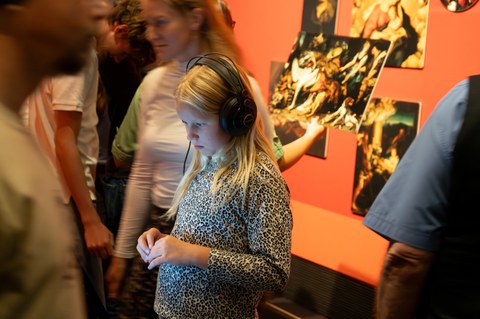 A girl is standing in a bustling exhibition hall wearing headphones. She is illuminated by a display.