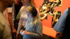 A girl is standing in a bustling exhibition hall wearing headphones. She is illuminated by a display.