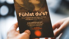 A hand is holding a dark flyer for the exhibition “Can You Feel It?”