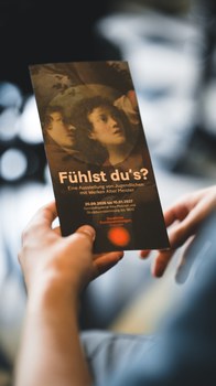 A hand is holding a dark flyer for the exhibition “Can You Feel It?”