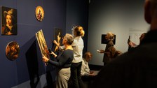 Two women are standing in front of a dark blue exhibition wall. One of them is holding one of the removable pictures.