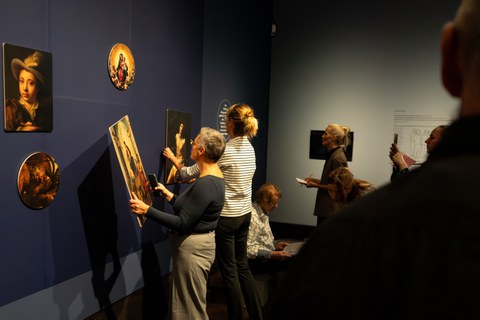 Two women are standing in front of a dark blue exhibition wall. One of them is holding one of the removable pictures.