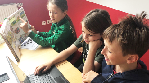 An einem Tisch sitzen drei Jungen und lernen. Einer zeigt den anderen zwein im Lehrbuch etwas. Die beiden Jungen im Vordergrund schreiben am Laptop. 