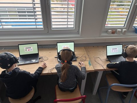 Three children are sitting with headphones on, each at a laptop with a computer mouse in their hand. There are texts and images on the screen.