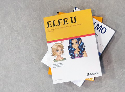 ELFE-material on top of a stack of folders containing research materials. On a yellow background is the title “ELFE 2. A reading comprehension test,” and below it, on a white background, are two manga-style comic characters.
