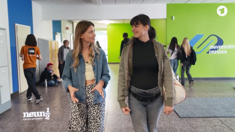 Two young women are walking down a school hallway and talking. In the background is a green wall with the logo of the Dresden University School.