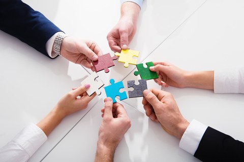 A bird's-eye view: Six hands are each holding a large colored puzzle piece, forming a circle.