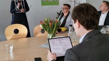 Schulleiterin Maxi Heß präsentiert in einem Besprechungsraum stehend vor drei sitzenden Männern. Kultusminister Clemens liest den Schulsteckbrief auf einem Tablet.