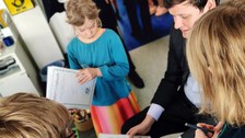 Drei Schüler:innen zeigen Kultusminister Conrad Clemens ihre Logbücher. Er sitzt auf einem Kinderstuhl und wird von ihnen umringt.