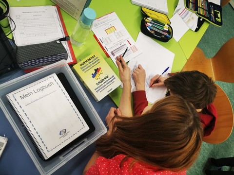 Vogelperspektive auf einen grünen Schreibtisch: Eine junge Frau hilft einem Schüler bei den Aufgaben. Auf dem Tisch liegen ein Logbuch der Universitätsschule Dresden und ein Deutsch-Ukrainisches Wörterbuch.
