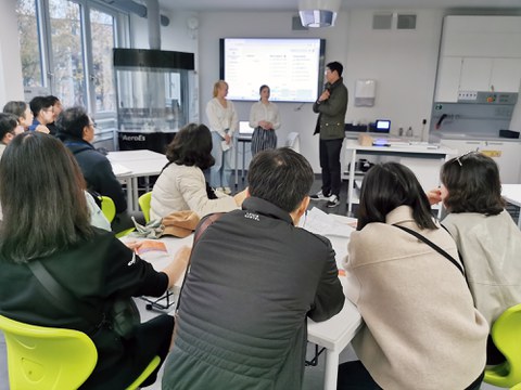 Two students and a translator stand in front of a Korean delegation in a classroom, in the background a large screen with a personalised timetable.