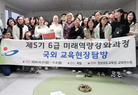 Two pupils and principal Maxi Heß stand surrounded by around 20 people from South Korea behind a banner reading ‘Improve future skills of Grades 5 and 6 - Educational trip through Germany - 27 Oct - 4 Nov 2024 - Gyeongsangnam-do Education Office