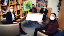 Im Rektorinnenbüro der Unievrsitätsschule Dresden sitzen Schulleiterin Maxi Heß, Elternrat Ulrich Puhlfürst, Mitglied des Universitätsschule Dresden e.V. Prof. Stefan Kornhuber und Prof. Anke Langner auf Sessel und Sofa um einen Sofatisch. Sie halten den übergroßen syambolischen Spendenscheck in die Kamera.