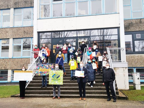 Auf der Treppe vor dem Schulgebäude steht die Delegation mit dem Spendenscheck, Million-Pixel-Bild und der Mappe mit den Unterschriften