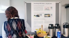 Eine Frau steht an einer Kaffeebar mit Früchten. Dahinter ein Poster "Forschung trifft Schule - Wie geht das?"