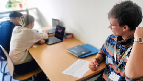 Ein junger Mann sitzt an einem Arbeitstisch und schaut auf zwei Schüler:innen. Sie besprechen sich am Laptop.