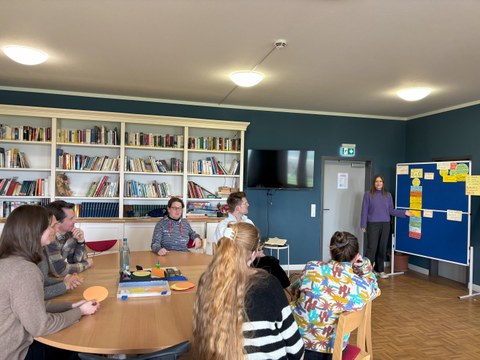 Foto. Ein Bibliotheksraum. An einem großen Tisch sitzen sieben Personen. Sie blicken zu einer Person, die an Wandtafel steht und gerade anhand von Notizzetteln etwas erklärt.