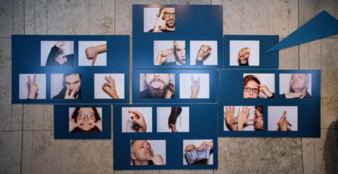 Foto der Ausstellung "Schmähung Provokation Stigma" 2020 in der SLUB, zu sehen mehrere Gesichter und Hände mit beleidigenden Gesten und Grimassen