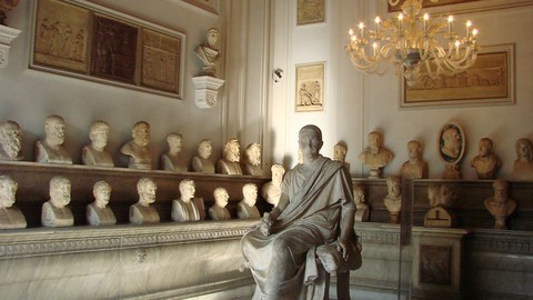 Saal in klassischer Architektur mit Büsten auf zwei umlaufenden Regalen, in der Mitte die Skulptur eines sitzenden Konsuls.