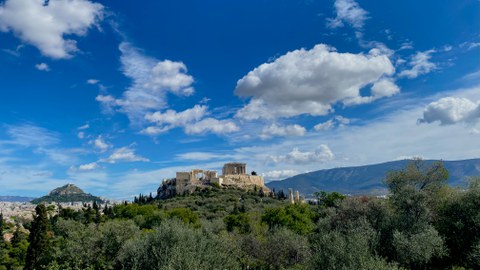 Panorama: zentral ist die Akropolis, links der Lykabettos-Berg. Ein sonniger Tag mit einzelnen Wolken. Im Hintergrund ein längliches Bergmassiv mit einem Windkraftwerk