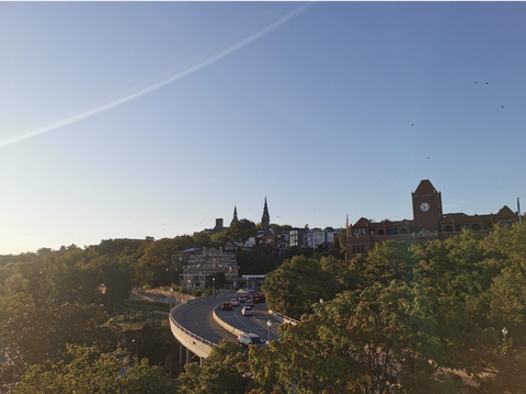 Das Bild zeigt die hinter Bäumen hervorragende Georgetown University in Washington DC aus der Ferne im Abendlicht, davor  eine breitspurig von Autos befahrene Brücke.