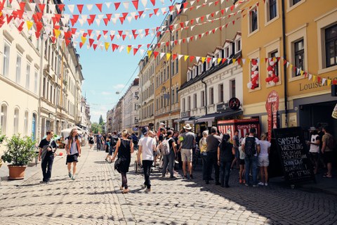 Zu sehen ist eine helle Straße, auf der Menschen laufen und stehen. Zwischen den Häusern links und rechts sind bunte Wipfel gespannt.