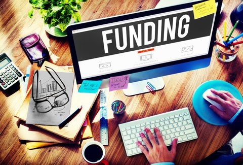Bild eines Arbeitsplatzes mit Computer. Auf dem Desktop steht groß das Wort "FUNDING".