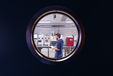 Blick durch ein rundes, bullaugenförmiges Fenster in ein Labor der Biotechnologie mit verschiedenen Messgeräten, durch das gerade ein junger bärtiger Forscher läuft.