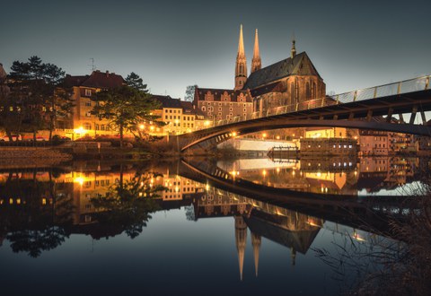 Blick über die Neisse auf die Altstadtbrücke und die abendlich erleuchtete Görlitzer Altstadt mit der prägnanten Silhouette der Peterskirche. Die beleuchtete Altstadt spiegelt sich im Fluss.