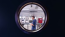 Blick durch ein rundes, bullaugenförmiges Fenster in ein Labor der Biotechnologie mit verschiedenen Messgeräten, durch das gerade ein junger bärtiger Forscher läuft.