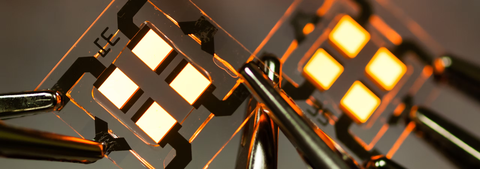 Vier quadratische, orange leuchtende Flächen, die sich auf einem größeren Glassubstrat befinden, einschließlich elektrischer Leiterbahnen und kontaktierten Krokodilklemmen.