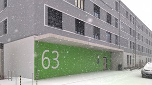 Das Bürogebäude N63 bei starkem Schneefall. Es ist ein moderner, graufarbiger Neubau mit einer großen grünen Fläche auf der die Hausnummer von weitem erkennbar ist.