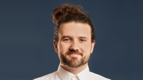 The person Ron Hildebrandt in a white suit top and a soft smile looking directly into the camera. He has brown eyes, long brown pinned up hair and wears a beard. The background is dark blue. 