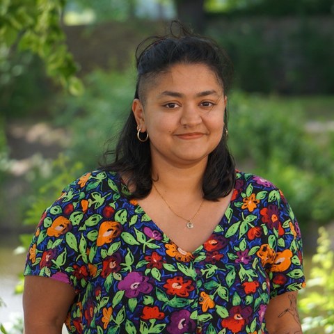 Nirbhika smiles into the camera. She wears a coloful dress with flowers. In the background green trees and plants can be seen.